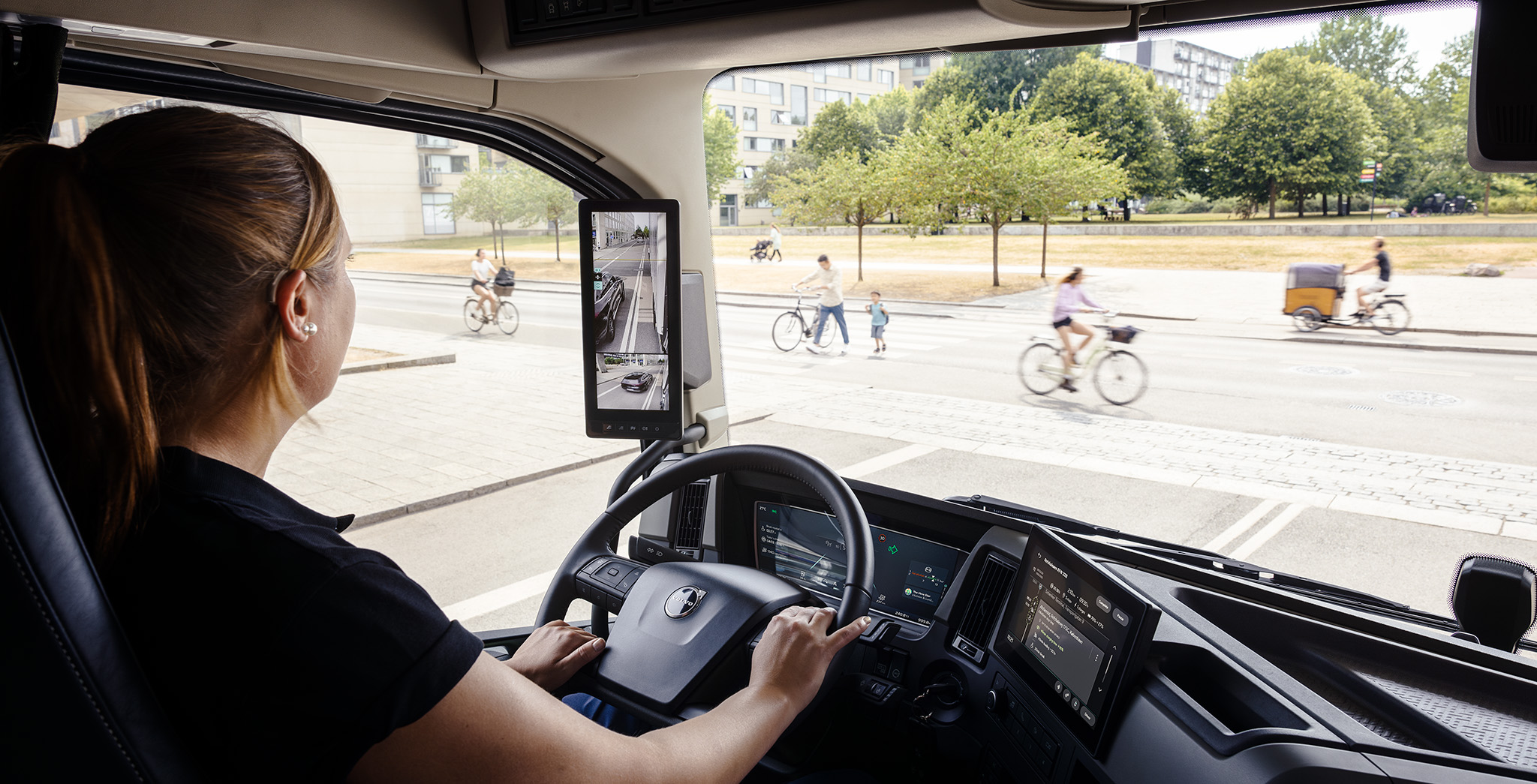 Sunkvežimio vairuotojas modernioje kabinoje, naudojant šonines kameras, važiuojant miesto gatve su dviratininkais ir pėstaisiais.