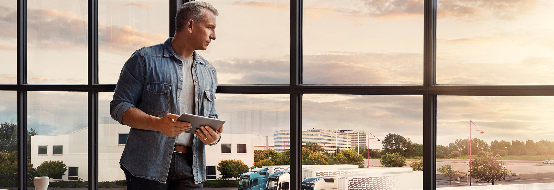 Planšetinį kompiuterį laikantis vyras stovi prie lango ir žvelgia žemyn į savo sunkvežimių parką